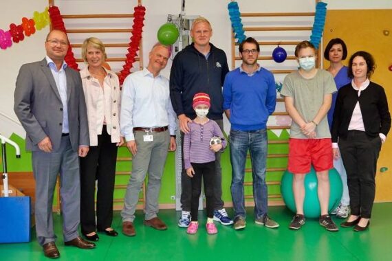 Eine Gruppe Erwachsener steht um ein Kind in einem mit bunten Luftballons und Wandmalereien geschmückten Krankenhausspielzimmer. Einige Personen lächeln, und zwei Personen tragen medizinische Masken.