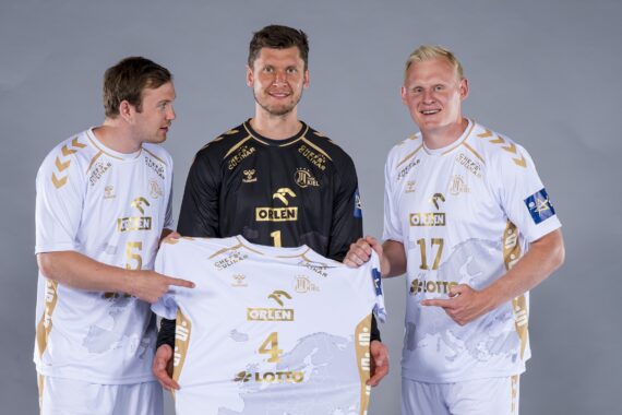 Drei männliche Sportler in weiß-goldenen Handballtrikots posieren für ein Foto; der Spieler in der Mitte, der ein schwarzes Torwarttrikot trägt, hält ein weißes Trikot mit der Nummer 4. Alle lächeln vor einem schlichten grauen Hintergrund.