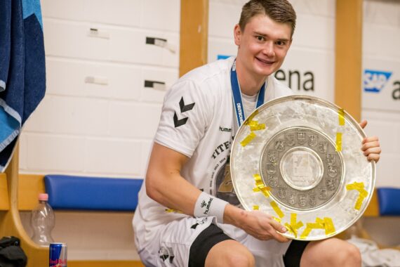 Ein lächelnder Sportler in weißer Uniform sitzt auf einer Bank in einer Umkleidekabine und hält eine große silberne Trophäenplatte, die mit gelbem Band verziert ist. Um seinen Hals hängen Medaillen, und in der Nähe sind Handtücher und Wasserflaschen zu sehen.