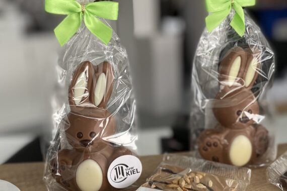 Zwei Schokoladenhasenfiguren, verpackt in durchsichtigem Plastik mit grünen Schleifen, auf einem Tisch ausgestellt. Auf der Verpackung befindet sich ein kleines Etikett mit der Aufschrift KIEL. Im Vordergrund sind auch Nüsse und Schokoladenstücke zu sehen.