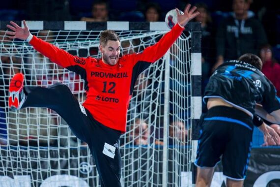 Ein Handballtorwart in einem roten Trikot mit ausgebreiteten Armen und Beinen blockiert einen Torschuss, während ein gegnerischer Spieler in Schwarz versucht, ein Tor zu erzielen. Die Szene ist dynamisch und actionreich.