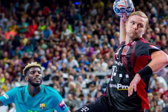 Ein Handballspieler in einem schwarz-roten Trikot bereitet sich auf den Wurf vor, während ein Verteidiger in einem blaugrünen Trikot zusieht. Die Aktion findet in einer vollbesetzten Arena mit einer unscharfen Zuschauermenge im Hintergrund statt.