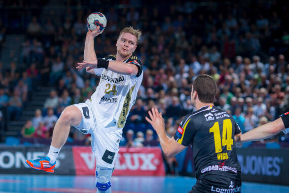 Ein männlicher Handballspieler in einem weißen Trikot springt, um den Ball zu werfen, während ein Verteidiger in einem schwarzen Trikot mit der Nummer 14 die Arme hebt, um ihn abzuwehren. Das Geschehen findet in einer Halle statt, in der eine Menschenmenge zuschaut.