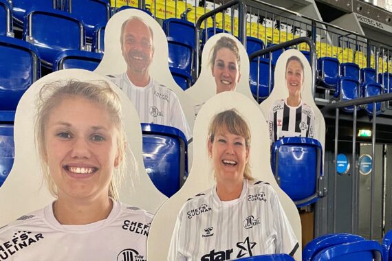 Pappfiguren von lächelnden Menschen in weißen Sporttrikots sind auf blauen Stadionsitzen platziert, um Fans auf der Tribüne zu simulieren. Der Bereich ist gut beleuchtet, im Hintergrund sind gelbe Sitze zu sehen.