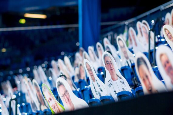 Auf den blauen Stadionsitzen sind reihenweise Pappausschnitte mit den Gesichtern von Menschen platziert, so dass der Anschein erweckt wird, dass das Publikum in einer ansonsten leeren Arena anwesend ist.