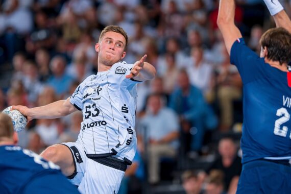 Ein Handballspieler in einem weißen Trikot springt und bereitet sich auf den Wurf vor, während zwei Gegner in blauen Trikots versuchen, ihn während eines intensiven Spiels in einer überfüllten Halle zu blockieren.