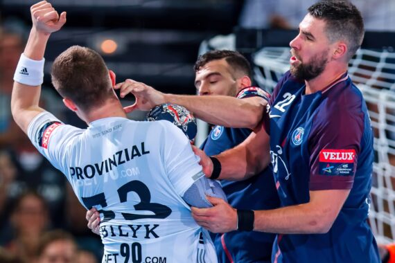 Zwei Handballspieler in blauen Trikots blockieren einen Spieler in einem weißen Trikot während eines intensiven Spiels. Ein Spieler im blauen Trikot ergreift den Arm des Spielers im weißen Trikot in der Nähe des Torraums.