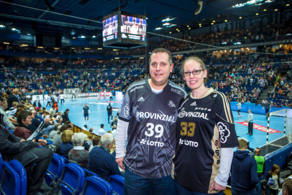 Ein lächelnder Mann und eine lächelnde Frau in passenden Handballtrikots stehen in einer Halle mit blauen Sitzen, umgeben von einer jubelnden Menge und einem Handballspiel auf dem Spielfeld im Hintergrund.