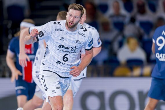 Ein männlicher Handballspieler in einem weißen Trikot mit der Nummer 6 läuft während eines Spiels auf dem Spielfeld, umgeben von Gegnern in blauen Trikots. Im unscharfen Hintergrund sind Zuschauer zu sehen.
