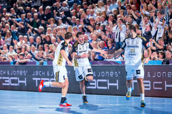 Drei männliche Handballspieler in weißen und schwarzen Trikots feiern auf einem Hallenplatz, während eine jubelnde Menge die Tribüne hinter ihnen füllt. Die Atmosphäre ist energiegeladen und lebendig.