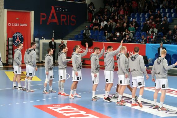 Eine Herrenhandballmannschaft in grauen Kapuzenpullis und weißen Shorts reiht sich vor einem Spiel auf einem Hallenplatz auf und gibt sich ein High-Five. Die Zuschauer sitzen auf blauen Sitzen, und die Halle ist mit Paris-Schildern geschmückt.