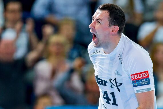 Ein männlicher Handballspieler in einem weißen Trikot schreit leidenschaftlich, wahrscheinlich beim Feiern während eines Spiels. Im Hintergrund ist eine unscharfe, jubelnde Menge zu sehen.