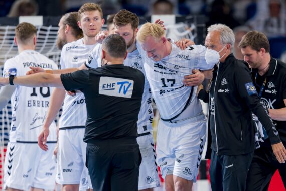Ein Handballspieler in einem weißen Trikot wird nach einer offensichtlichen Verletzung von Mannschaftskameraden und Betreuern vom Spielfeld getragen, während ein Schiedsrichter in der Nähe steht. Andere Spieler schauen mit Sorge zu.
