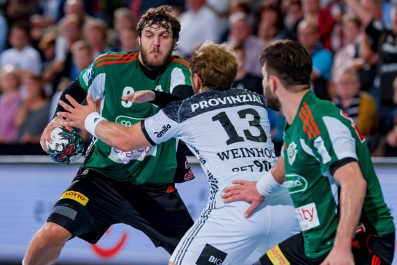 Ein Handballspieler in einem grün-schwarzen Trikot hält den Ball, während er von zwei Spielern in weiß-schwarzen Trikots während eines intensiven Spiels verteidigt wird, während im Hintergrund eine Menschenmenge zu sehen ist.
