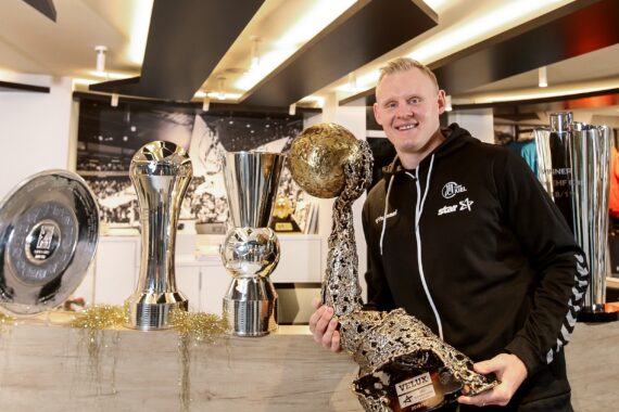 Ein lächelnder Mann steht im Haus und hält eine große, strukturierte goldene Sporttrophäe in der Hand. Auf der Theke neben ihm stehen mehrere andere glänzende Trophäen in verschiedenen Formen und Größen.