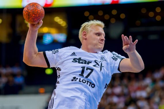 Ein männlicher Handballspieler in einem weißen Trikot mit der Nummer 17 bereitet sich darauf vor, während eines Spiels einen orangefarbenen Ball zu werfen, während im Hintergrund eine unscharfe Menschenmenge zu sehen ist.