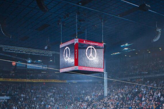 Eine große Anzeigetafel in der Arena zeigt ein Bild des Eiffelturms in Form eines Friedenssymbols. Die Arena ist mit einer stehenden Menge gefüllt, und die Atmosphäre wirkt feierlich und respektvoll.