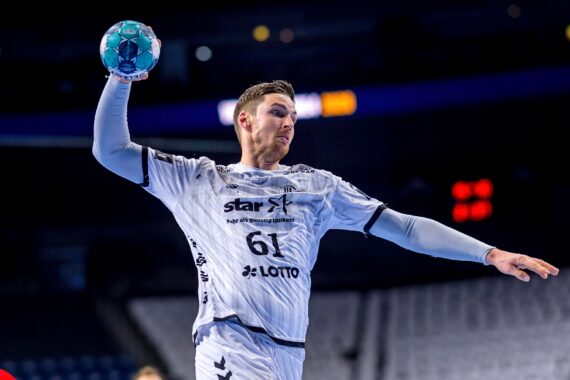 Ein männlicher Handballspieler in einem weißen Trikot mit der Nummer 61 springt und bereitet sich darauf vor, den Ball während eines Spiels in einer Halle mit leeren Sitzen im Hintergrund zu werfen.