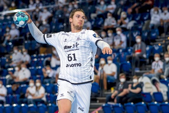 Ein Handballspieler in einem weißen Trikot springt in die Luft, hält einen blauen Ball und bereitet sich auf den Wurf vor. Maskierte Zuschauer schauen von blauen Stadionsitzen im Hintergrund zu.
