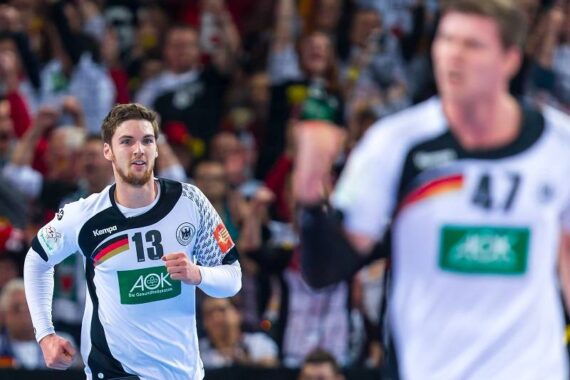 Zwei männliche Handballspieler in weißen und schwarzen Deutschland-Trikots befinden sich auf einem Spielfeld, wobei einer (Nummer 13) im Fokus läuft und der andere (Nummer 47) unscharf im Vordergrund steht. Im Hintergrund sind Zuschauer zu sehen.