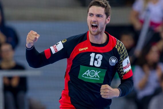 Ein männlicher Handballspieler, der ein schwarz-rotes Trikot mit der Nummer 13 trägt, jubelt mit geballter Faust und aufgeregtem Gesichtsausdruck während eines Spiels.