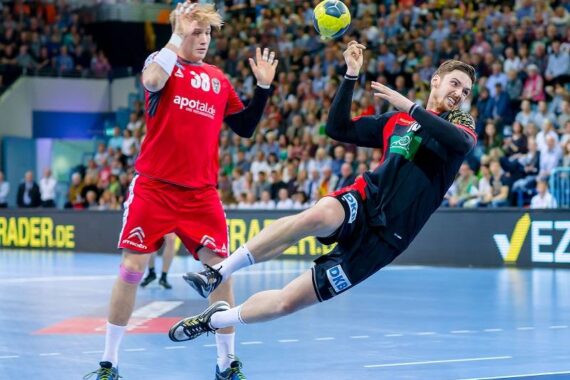 Ein Handballspieler in einem schwarzen Trikot springt zur Seite, um den Ball zu schießen, während ein Verteidiger in Rot die Arme zum Abwehren hebt. Eine große Menschenmenge verfolgt das Geschehen in der Halle.