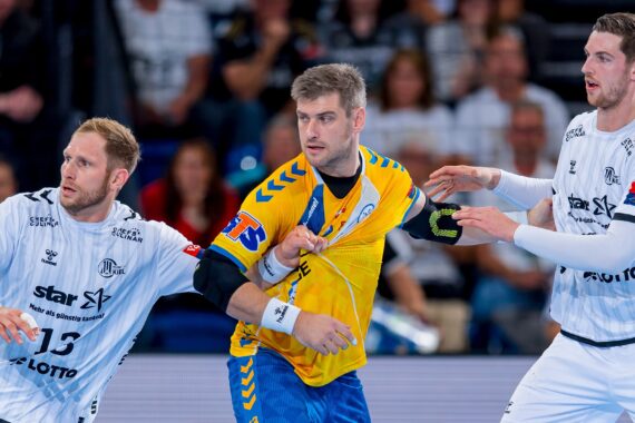 Drei männliche Handballspieler in Aktion während eines Spiels. Ein Spieler in einer gelb-blauen Uniform hält den Ball, während zwei Spieler in weißen Uniformen versuchen, ihn zu verteidigen. Die Szene spielt sich in einer Halle ab, mit Zuschauern im Hintergrund.