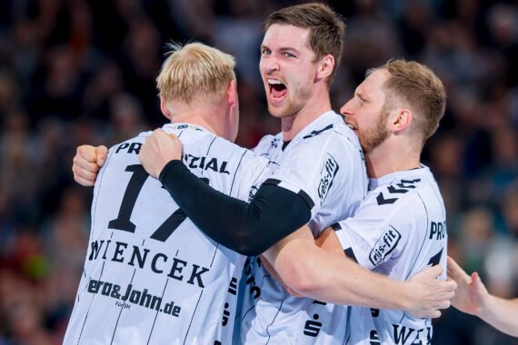 Drei männliche Handballspieler in weißen Trikots feiern zusammen, lächeln und umarmen sich auf dem Spielfeld während eines Spiels, mit einer unscharfen Menschenmenge im Hintergrund.