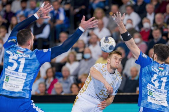 Ein Handballspieler in einem weißen Trikot wirft den Ball, während zwei Verteidiger in blauen Trikots mit erhobenen Armen versuchen, ihn abzublocken, während die Zuschauer im Hintergrund zuschauen.