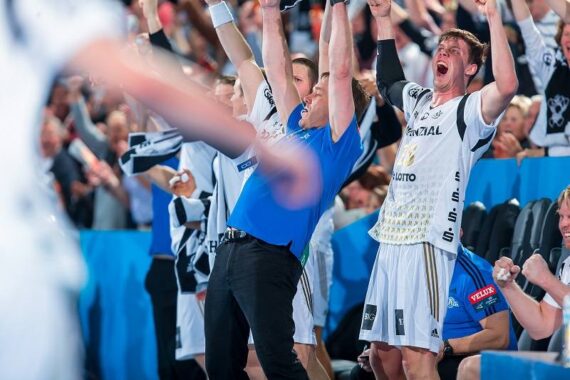 Eine Gruppe ekstatischer Handballspieler und ein Trainer feiern mit erhobenen Armen und jubelnd, was Aufregung und Freude während eines Spiels zeigt. Der Hintergrund ist mit begeisterten Fans gefüllt.