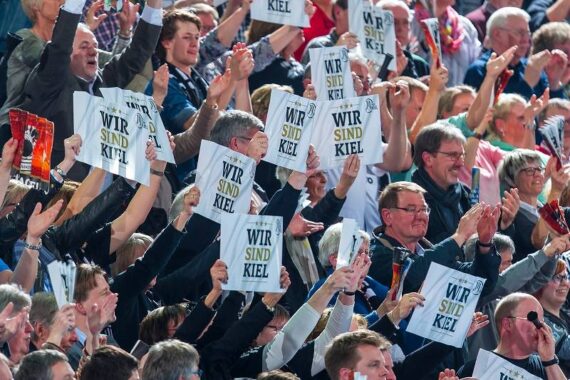 Eine große Menschenmenge jubelt und hält Schilder mit der Aufschrift WIR SIND KIEL bei einer Veranstaltung im Freien hoch, um Begeisterung und Einigkeit zu zeigen. Viele lächeln und heben ihre Arme in die Luft.