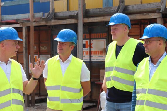 Vier Männer mit blauen Schutzhelmen und gelben Sicherheitswesten stehen auf einer Baustelle. Ein Mann spricht und gestikuliert mit seinen Händen, während die anderen aufmerksam zuhören. Im Hintergrund sind Gerüste und Baumaterialien zu sehen.