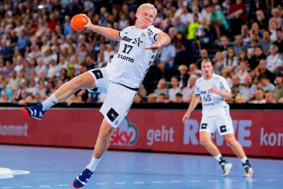 Ein Handballspieler in einem weißen Trikot springt in der Luft, um den Ball während eines Spiels zu werfen, während im Hintergrund ein anderer Spieler und eine dicht gedrängte Menge zu sehen sind.