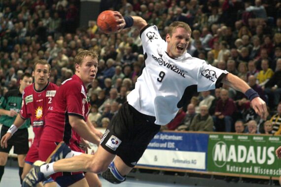 Ein Handballspieler in einem weißen Trikot springt mit dem Ball in der Hand in die Luft, während ein Verteidiger in einem roten Trikot zuschaut. Das Spiel findet in einer mit Zuschauern gefüllten Halle statt.