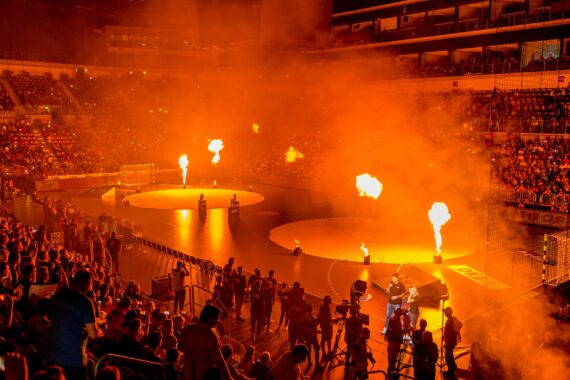 Eine große überdachte Arena, gefüllt mit Zuschauern, die ein dramatisches Ereignis verfolgen, mit Feuer- und Raucheffekten, die das Hauptgeschoss erhellen. Die Atmosphäre ist energiegeladen und lebendig, mit leuchtend orangefarbenen Flammen, die sich von der dunklen Umgebung abheben.