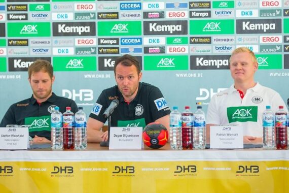Drei Männer sitzen an einem Pressekonferenztisch vor einer Kulisse mit Sponsorenlogos. Der Mann in der Mitte spricht in ein Mikrofon, während die beiden anderen auf beiden Seiten sitzen, mit Mikrofonen, Namensschildern und Wasserflaschen vor sich.