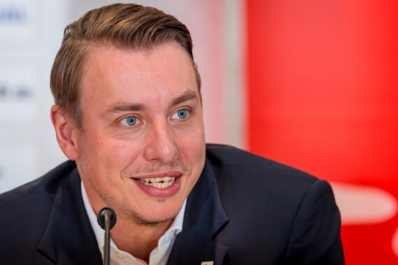 Ein Mann in einem dunklen Anzug und weißem Hemd lächelt, während er während einer Pressekonferenz in ein Mikrofon spricht, mit einem verschwommenen roten und weißen Hintergrund hinter ihm.