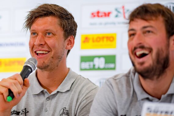 Zwei Männer in grauen Poloshirts lächeln und sprechen auf einer Pressekonferenz. Einer hält ein Mikrofon in der Hand, während auf einem Hintergrund hinter ihnen Sponsorenlogos zu sehen sind.