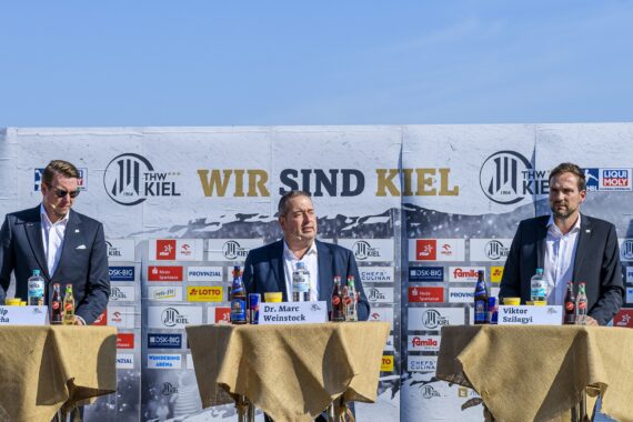 Drei Männer in Anzügen stehen bei einer Presseveranstaltung im Freien hinter separaten, mit beigen Tüchern bedeckten Tischen, mit Mikrofonen und Getränkeflaschen vor einem Hintergrund mit der Aufschrift WIR SIND KIEL.