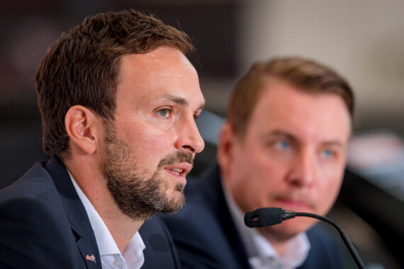 Zwei Männer in Anzügen sitzen an einem Tisch mit Mikrofonen und sprechen oder hören während einer Konferenz oder Besprechung zu. Der Mann im Vordergrund ist scharf, während der Mann im Hintergrund unscharf ist.