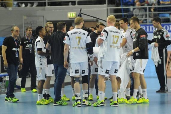 Eine Herren-Handballmannschaft in weißen Trikots und neongelben Schuhen versammelt sich während eines Spiels mit den Trainern auf einem Hallenplatz zu einem Gedränge. Im Hintergrund sind Zuschauer zu sehen.