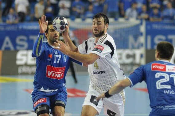 Drei männliche Handballspieler treten während eines Spiels gegeneinander an; ein Spieler in einem weißen Trikot hält den Ball, während zwei Spieler in Blau versuchen, ihn zu blockieren. Im unscharfen Hintergrund sind Zuschauer und Fahnen zu sehen.