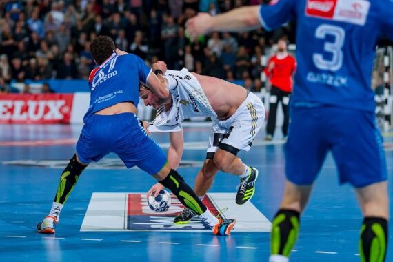 Zwei männliche Handballspieler kämpfen auf einem blauen Spielfeld um den Ball; der eine in einer blauen Uniform packt das Hemd des anderen, der sich in einer weißen Uniform nach vorne beugt. Die Szene ist intensiv, mit Zuschauern im Hintergrund.