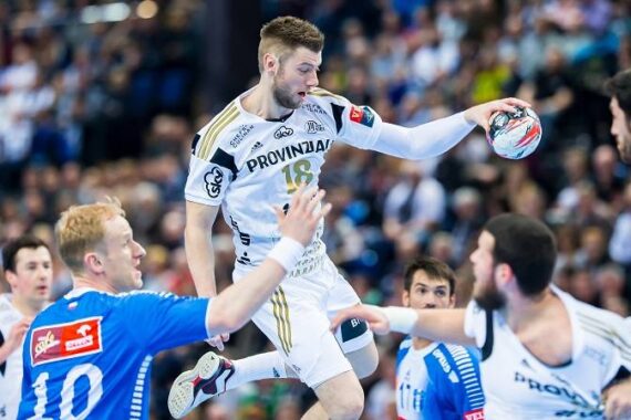 Ein Handballspieler in einem weißen Trikot springt in der Luft, um den Ball zu werfen, während Spieler in blauen Trikots versuchen, ihn während eines intensiven Spiels zu verteidigen. Im Hintergrund sind Zuschauer zu sehen.
