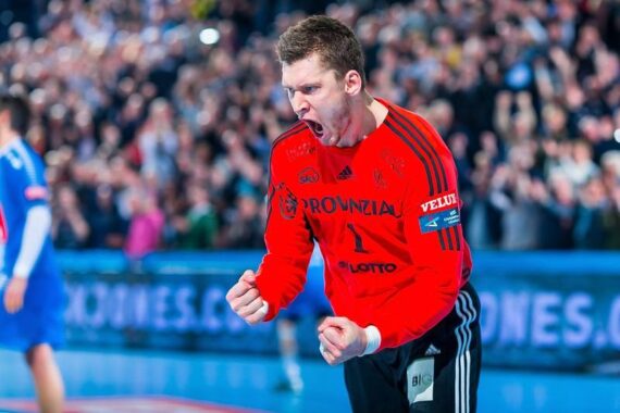 Ein männlicher Handballspieler in einem roten Trikot ballt die Fäuste und jubelt auf einem Hallenplatz, während im Hintergrund eine jubelnde Menge zu sehen ist.