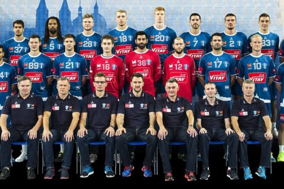 Eine Herrenhandballmannschaft und ihre Trainer posieren gemeinsam für ein Gruppenfoto. Die Spieler in blauen und roten Trikots stehen hinter den Trainern in schwarzen Poloshirts und Hosen vor einem blauen Hintergrund mit einem Stadtmotiv.