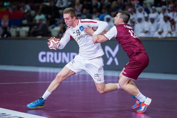 Ein Handballspieler in einem weißen Trikot läuft mit dem Ball, während er von einem Gegner in einem kastanienbraunen Trikot auf einem Hallenplatz eng verteidigt wird, während die Zuschauer im Hintergrund zusehen.