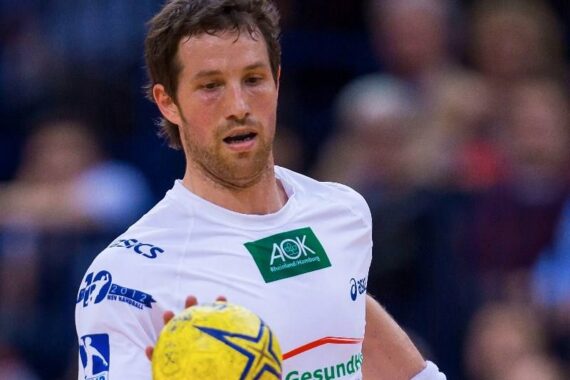 Ein männlicher Handballspieler in einem weißen Trikot konzentriert sich auf einen gelben Handball, während er in einer Halle spielt. Der Hintergrund ist unscharf und hebt das Geschehen hervor.