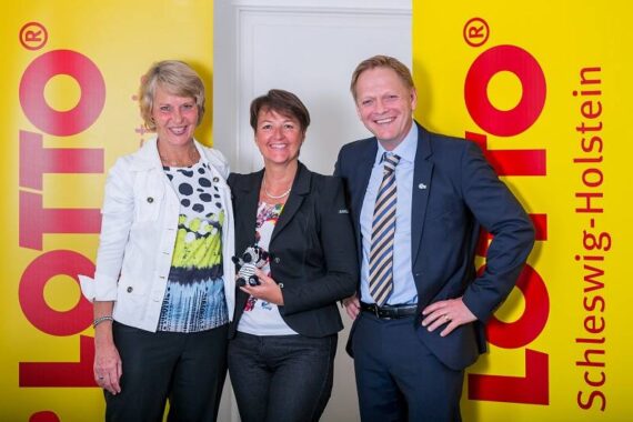 Drei Personen in Business-Kleidung stehen lächelnd vor gelben Bannern mit der Aufschrift LOTTO Schleswig-Holstein. Die beiden Frauen stehen auf der linken Seite, der Mann auf der rechten. Die Frau in der Mitte hält einen kleinen Pokal in der Hand.