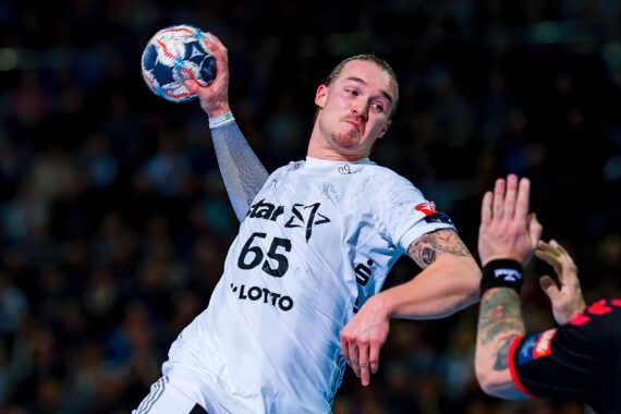 Ein Handballspieler im weißen Trikot mit der Nummer 65 springt mit dem Ball in der Hand zum Wurf, während ein Gegner im schwarzen Trikot während eines intensiven Spiels die Hand zum Blocken hebt.
